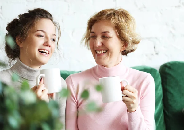 Due belle donne- madre e figlia sedute sul divano di casa. Bevono caffè, tè . — Foto Stock