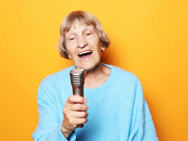 Feliz anciana cantando con micrófono, divirtiéndose, expresando talento musical sobre fondo amarillo — Foto de Stock