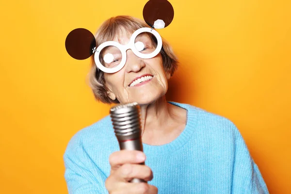 Mulher velha feliz com óculos grandes segurando um microfone e cantando isolado no fundo amarelo — Fotografia de Stock