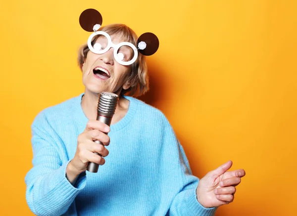 Ευτυχισμένη ηλικιωμένη γυναίκα με μεγάλα γυαλιά που κρατάει ένα μικρόφωνο και τραγουδάει απομονωμένη σε κίτρινο φόντο — Φωτογραφία Αρχείου