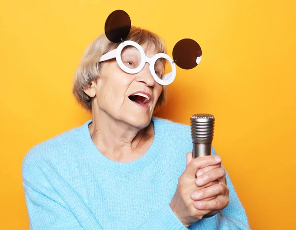 Szczęśliwa staruszka z dużymi okularami trzymająca mikrofon i śpiewająca na żółtym tle — Zdjęcie stockowe