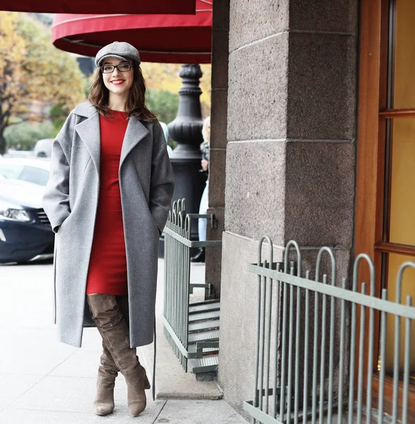 Jovem elegante mulher bonita vestindo vestido vermelho, casaco cinza e chapéu posando nas ruas da cidade . — Fotografia de Stock