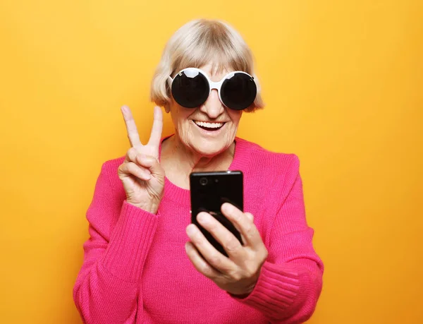 Ηλικιωμένη κρατάει ένα smartphone και καθιστώντας v-sign — Φωτογραφία Αρχείου