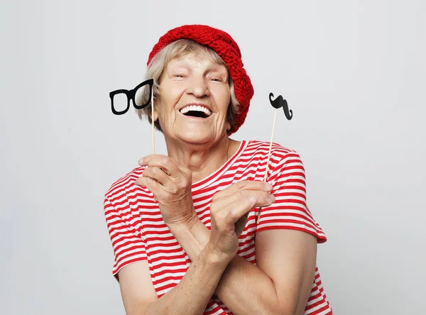 Engraçada avó com bigode falso e óculos, ri e se prepara para a festa sobre fundo cinza — Fotografia de Stock