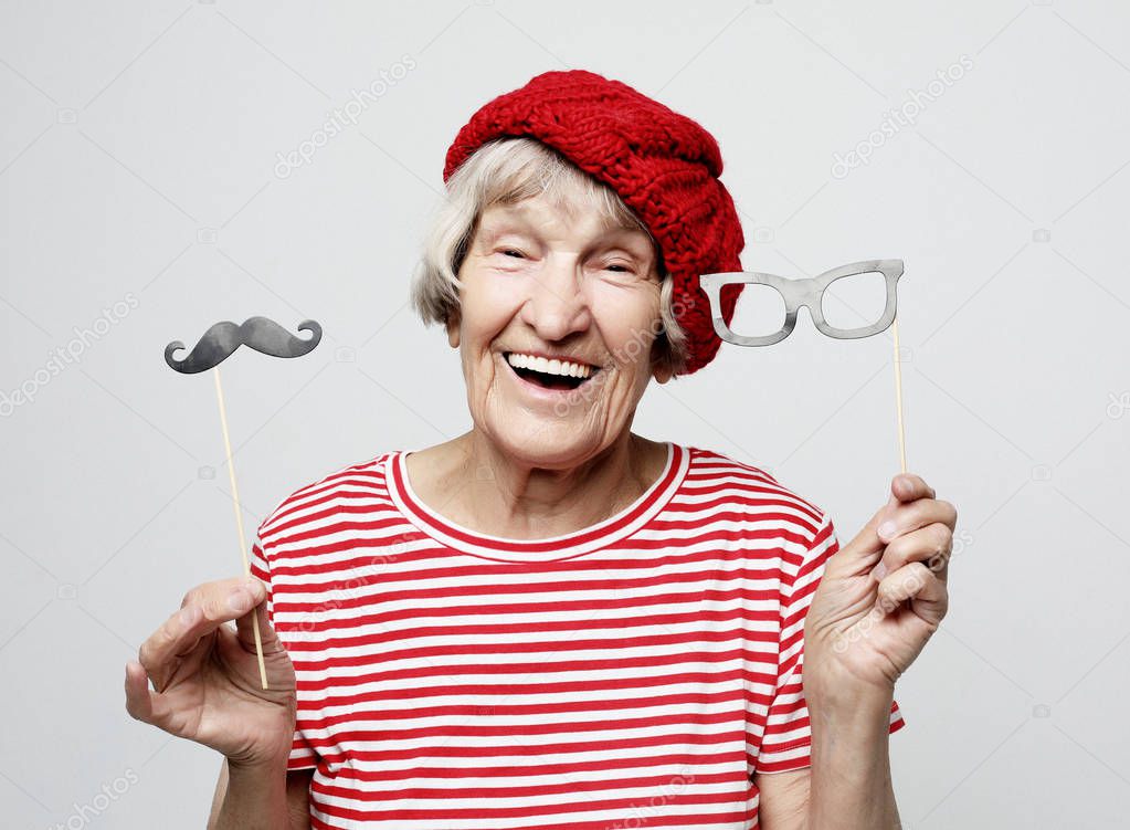 funny grandmother with fake mustache and glasses, laughs and prepares for party over grey background