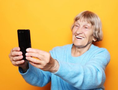 Mavi kazaklı büyükanne gülümsüyor ve sarı arka planda selfie çekiyor.