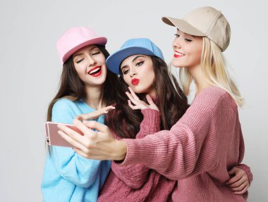 Üç mutlu en iyi kız arkadaş kazak ve şapka giyip akıllı telefondan selfie çekiyorlar..