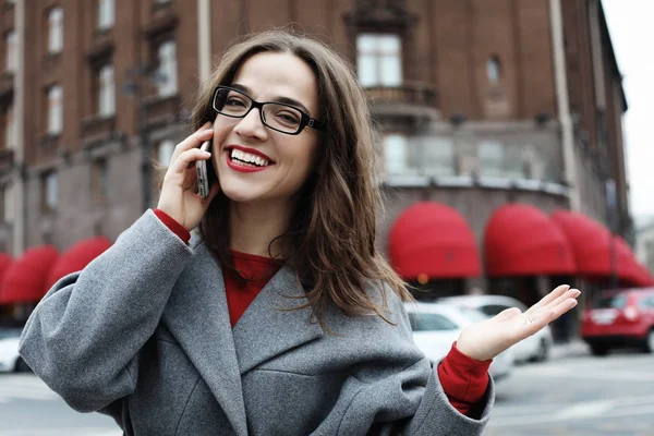 Retrato al aire libre de una joven sorprendida con gafas con abrigo gris y smartphone en la mano . — Foto de Stock