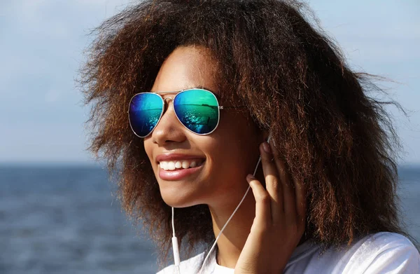 Encantador incrível afro-americano jovem mulher em óculos de sol ouvir música em fones de ouvido em seu telefone móvel . — Fotografia de Stock