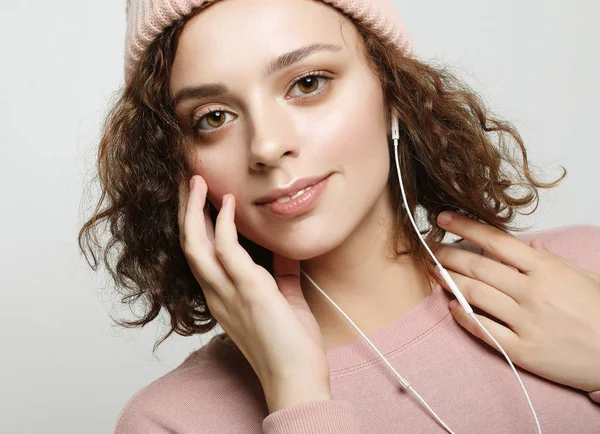 Beautiful girl in headphones listens to music and smiles while standing on a white background in a pink sweatshirt and hat. — Stock Photo, Image