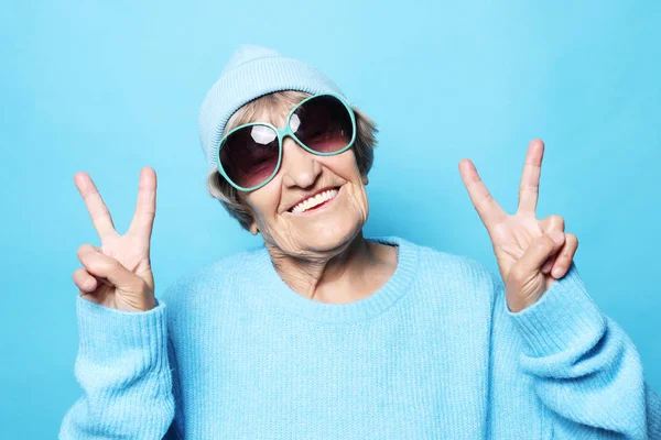 Estilo de vida, emoción y concepto de la gente: Señora de edad divertida con suéter azul, sombrero y gafas de sol que muestran signo de victoria. — Foto de Stock