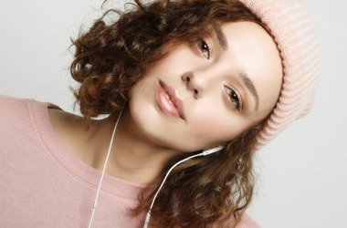 Kulaklıklı güzel bir kızın portresi pembe bir kazak ve şapkayla beyaz bir arka planda dururken müzik dinliyor ve gülümsüyor. Yaşam tarzı konsepti. Kapat..