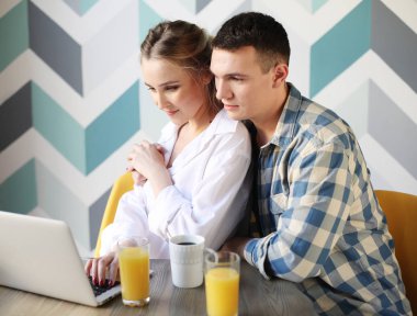 Kahkahalarla gülen bir çift kahvaltı yaparken mutfaktaki masada oturup dizüstü bilgisayara bakıyor.