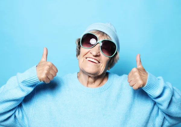 Yaşam tarzı, duygu ve insanlar konsepti: Mavi kazak giyen komik yaşlı kadın, şapka ve güneş gözlüğü zafer işareti gösteriyor. — Stok fotoğraf