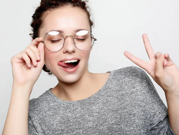 Giovane donna riccia con gli occhiali vestiti con disinvoltura facendo facce alla macchina fotografica, lampeggiando, sporgendo la lingua . — Foto Stock