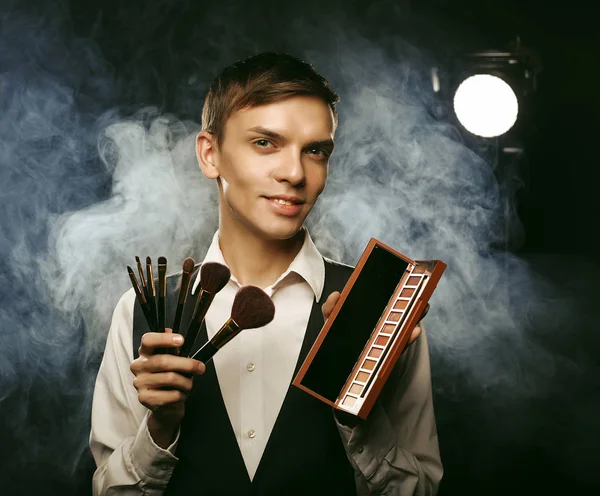 Young male make up artist posing with make-up brushes and shadow palette on a dark background. — Stock Photo, Image