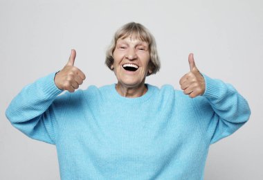 Yaşlı kadın başparmağını kaldırıyor ve kameraya bakıyor. 