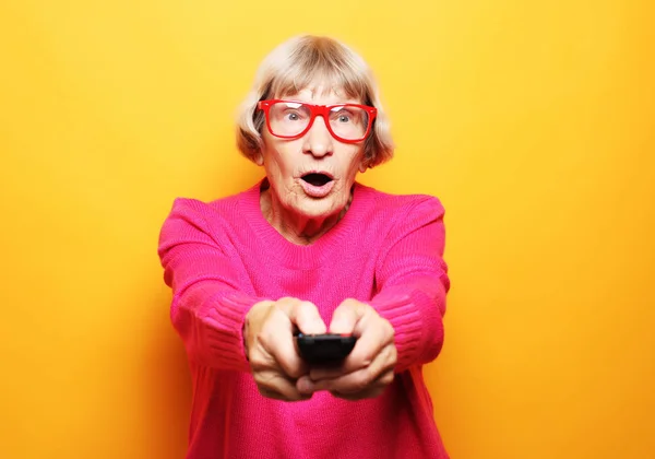 Lifestyle και άνθρωποι έννοια: αστεία γιαγιά κρατά ένα τηλεχειριστήριο τηλεόραση πάνω από κίτρινο φόντο — Φωτογραφία Αρχείου