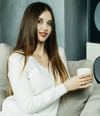 Güzel ve sevimli. Elinde kahve fincanı tutan ve evdeki sandalyeye oturan güzel genç bir kadın. Yaşam tarzı kavramı.