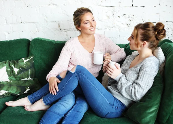 Duas mulheres lindas mãe e filha sentadas no sofá em casa. Eles bebem café, chá . — Fotografia de Stock