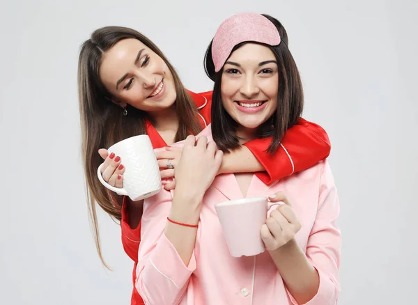 Två vackra flickor klädd i pyjamas kramas och hålla koppar — Stockfoto