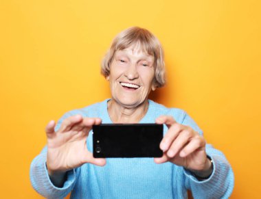 Yaşam tarzı, tehnoloji ve insan konsepti: büyükanne gülümser ve sarı arka planda selfie çeker