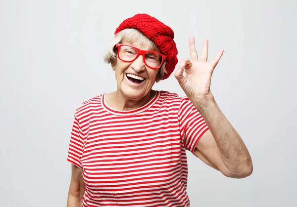 Estilo de vida, emoción y concepto de la gente: Divertida anciana con sombrero rojo y gafas que muestran signo ok — Foto de Stock