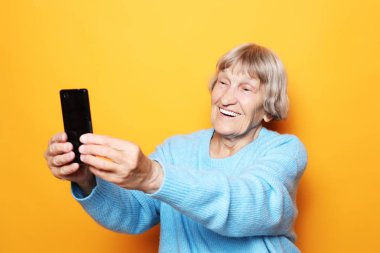Yaşam tarzı, tehnoloji ve insan konsepti: büyükanne gülümser ve sarı arka planda selfie çeker