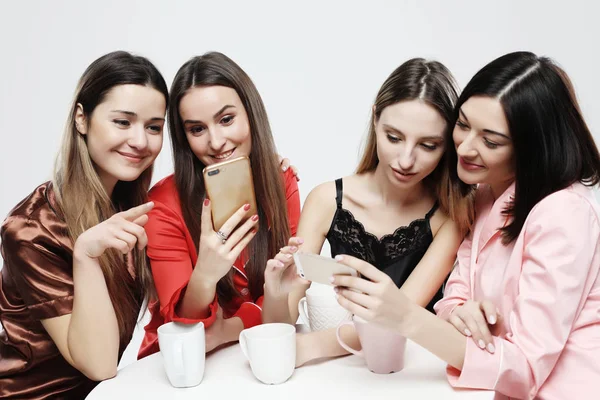 Cztery piękne koleżanki w piżamie siedzą przy stole, picie herbaty lub kawy i rozmowy w sieci społecznej. — Zdjęcie stockowe