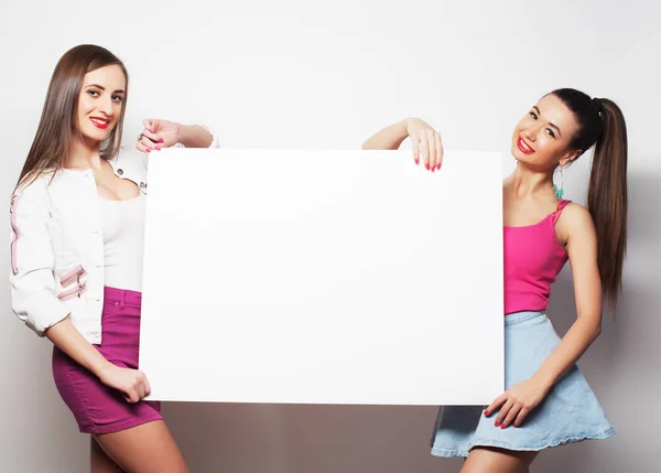 Two young hipster girl friends standing together and holding a blank billboard . Looking at camera Stock Image