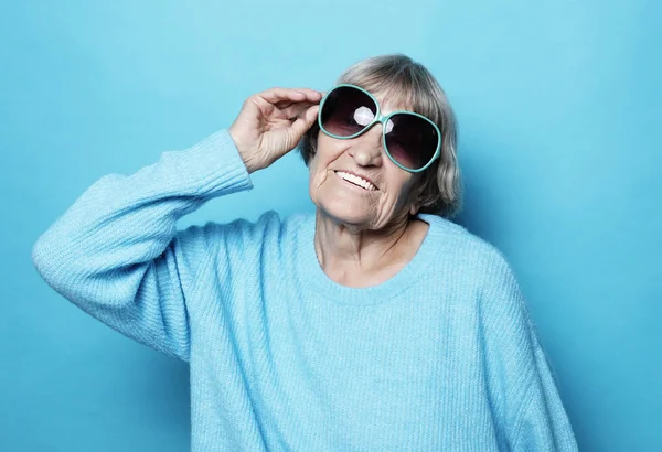 Životní styl, emoce a koncepce lidí: legrační stará dáma nosí modrý svetr a sluneční brýle. Izolované na modrém pozadí. — Stock fotografie
