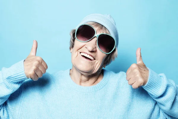 Grappige oude dame draagt blauwe trui, hoed en zonnebril overwinning teken tonen. Geïsoleerd op blauwe achtergrond. — Stockfoto