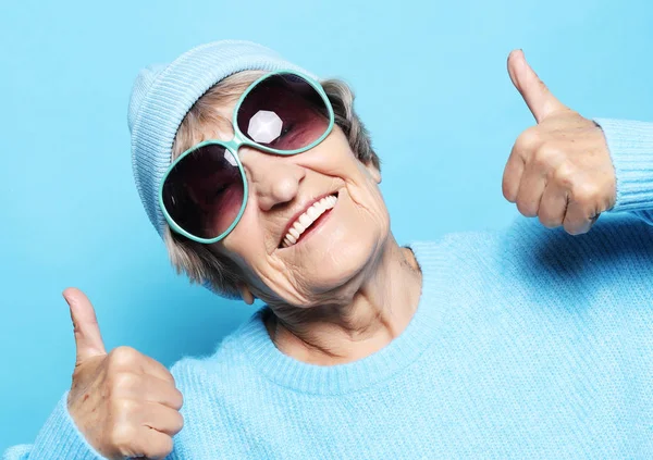 Lifestyle, emotion and people concept: Funny old lady wearing blue sweater, hat and sunglasses showing victory sign. — Stock Photo, Image