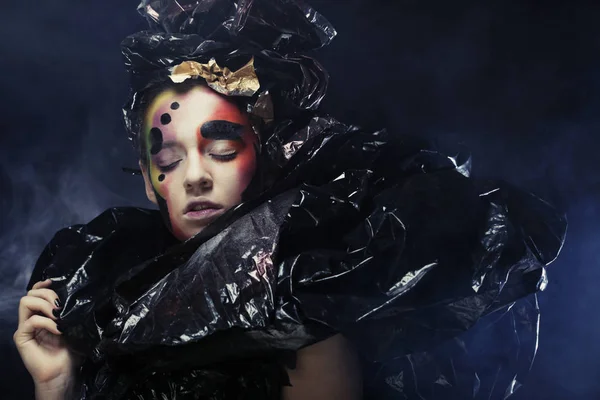 Hermosa joven con brillante fantasía maquillaje y traje posando sobre un fondo negro en las nubes de humo. Halloween — Foto de Stock