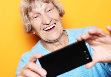 Mavi kazaklı büyükanne gülümsüyor ve sarı arka planda selfie çekiyor.