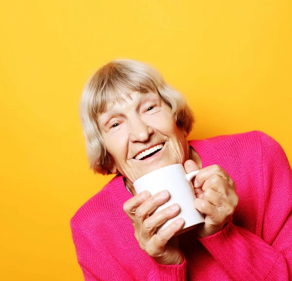 Portrait of old excited lady smiling laughing, holding cup drinking coffee, tea, beverage on yellow background