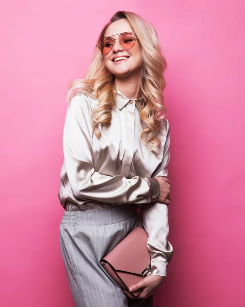 Junge schöne blonde Frau mit Bluse, Hose und Sonnenbrille hält eine Handtasche und posiert auf rosa Hintergrund. — Stockfoto