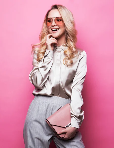 Junge schöne blonde Frau mit Bluse, Hose und Sonnenbrille hält eine Handtasche und posiert auf rosa Hintergrund. — Stockfoto