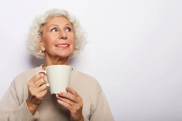 Velha senhora animada sorrindo rindo, segurando xícara bebendo café, chá, bebida no fundo cinza — Fotografia de Stock