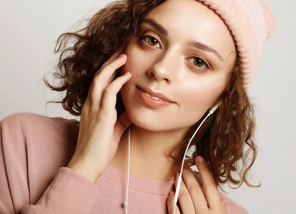 Ritratto di una bella ragazza in cuffia ascolta musica e sorride in piedi su uno sfondo bianco in felpa rosa e cappello . — Foto Stock