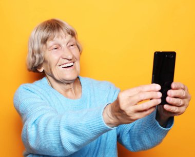 Mavi kazaklı büyükanne gülümsüyor ve sarı arka planda selfie çekiyor.