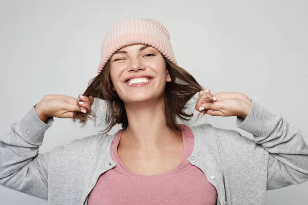 Mujer joven divertida en un sombrero de gorro rosa hace caras y posa sobre fondo gris claro — Foto de Stock