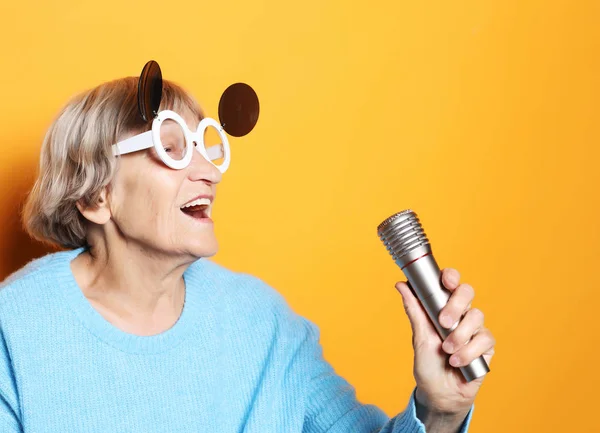 Boldog idős nő nagy szemüveg kezében egy mikrofon és énekel elszigetelt sárga háttér — Stock Fotó