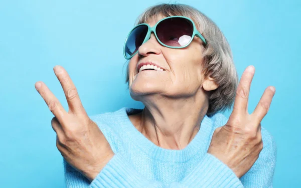 Legrační stará dáma na sobě modrý svetr, čepice a brýle zobrazující znamení vítězství — Stock fotografie