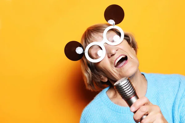 happy old woman with big eyeglasses holding a microphone and singing isolated on yellow background