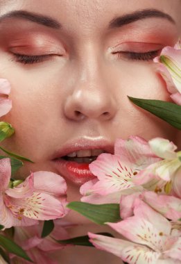 Güzel, genç bir kadın. Makyajı yumuşak renklerde. Kocaman pembe bir çiçek tutuyor.