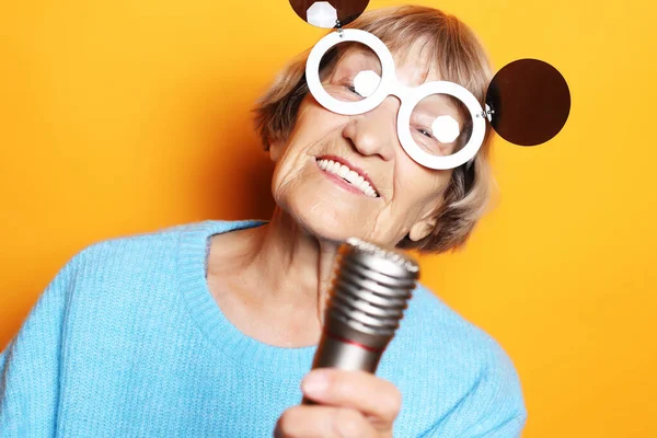 Mulher velha feliz com óculos grandes segurando um microfone e cantando isolado no fundo amarelo — Fotografia de Stock