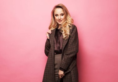 Güzel sarışın kadın pembe arka planda şık kıyafetler içinde poz veriyor.