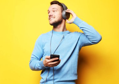 Yakışıklı genç adam, mavi kazak giyiyor, sarı arka planda müzik dinliyor.
