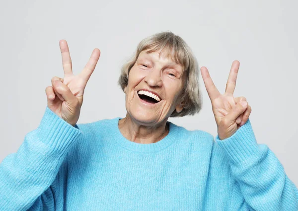 Gammal kvinna skrattar och visar fred eller seger signat kamera. Känslor och känslor. Porträtt av uttrycksfull mormor. — Stockfoto